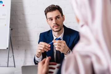serious translator holding pen while looking at arabian businesswoman on blurred foreground