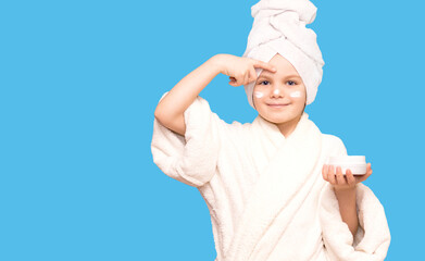 Caucasian child girl model with towel on her head puts cream on her face touching under eyes isolated on blue background. Skin care concept