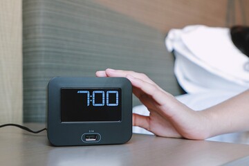 Woman hand turning off black digital alarm clock on the table side bed after awakening in morning...