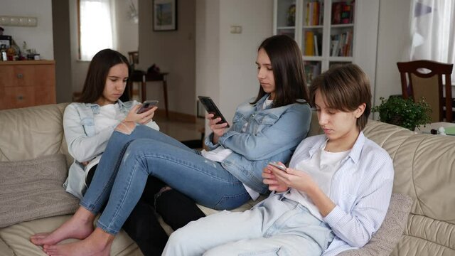 Teenager girls triplet sisters at home watch smart phones in social media sitting on couch