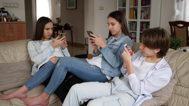 Teenager girls triplet sisters at home watch smart phones in social media sitting on couch