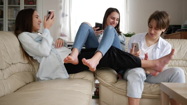 Teenager girls triplet sisters at home watch smart phones funny fighting with legs on couch