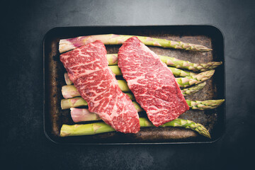Raw Steak with green asparagus on tray on black background, top view