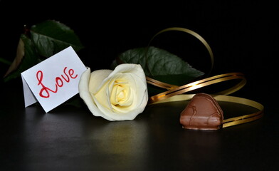 The studio white rose with golden ribbon on a black background. White leaflate with inscription love. Chocolate candy.