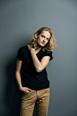 Young attractive guy with long blonde hair is sad leaning against a gray wall, looks to the camera
