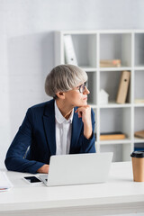 pensive team leader in glasses looking away near gadgets and coffee to go on desk