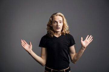 Young guy with long blond hair shows a gesture of perplexity, spreading his hands aside on a gray background