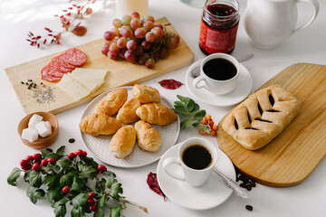 Continental breakfast of mini croissants, pastry, coffee in cups, grapes and jam in a jar, served on white table, decorated with white flowers bouquet, candles and sprig with green leaves