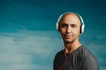 A bald man listens music in large headphones, Looks in surprise at the camera on a blue background