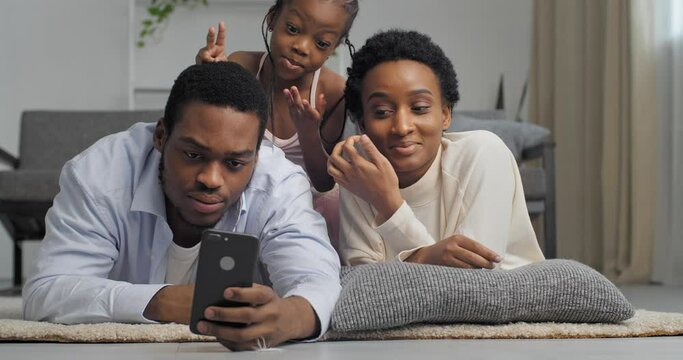 Three people, African American family with child girl lie on couch look at screen of mobile smartphone shoot video for blog, communicate via online chat conferences, take photo for camera pose smile