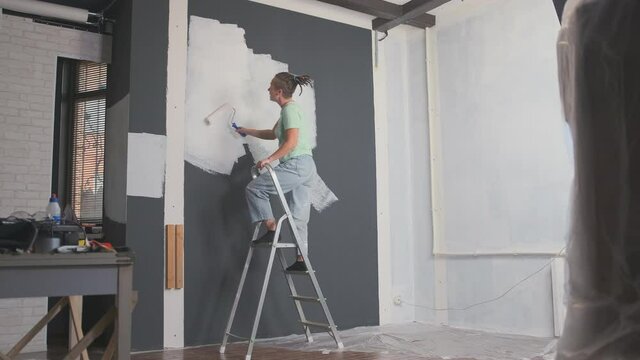 stylish designer or painter sings and dances while painting walls with paint roller in room standing on stepladder.
