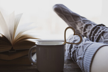 Lonely Valentine's Day. Love at distance, loneliness in self-isolation in the time of coronavirus. Selective focus on heart. Woman resting keeping legs in warm socks with morning coffee and books