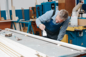 An elderly cabinetmaker in overalls and glasses paints a wooden board with a brush on a workbench...