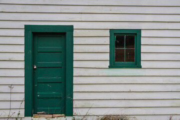 The side of an old shed with white siding and a green window and door.