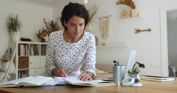 Concentrated smart millennial woman standing at table fill of different books, reading educational material, writing essay or preparing alone for exams, upgrading improving knowledge alone indoors.