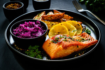 Fried salmon fillet with fried potatoes, red cabbage and lemon served on black plate on wooden table