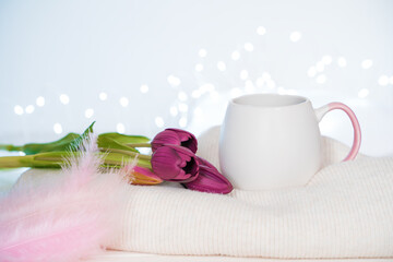 A bouquet of tulips, a mug of coffee, pink feathers and a sweater on a light background with burning lights. Side view, with space to copy. The concept of holiday backgrounds.