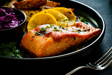 Fried salmon fillet with fried potatoes, red cabbage and lemon served on black plate on wooden table