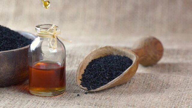 Glass bottle of black cumin seed essential oil , Nigella Sativa in scoop on black wooden background, oil dripping