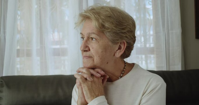 An elderly woman after retirement cut short hair with light brown hair is holding two hands on his chin and contemplating, distracted, feeling lonely at her home alone.