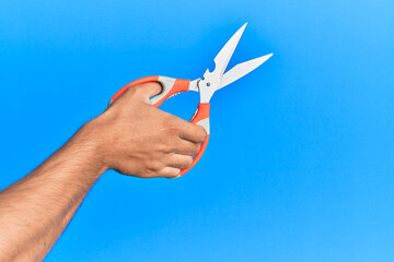 Hand of young hispanic man using scissors over isolated blue background.