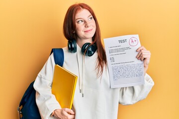Young red head girl wearing student backpack holding passed test smiling looking to the side and...