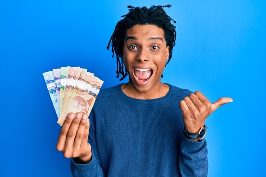 Young african american man holding canadian dollars pointing thumb up to the side smiling happy with open mouth