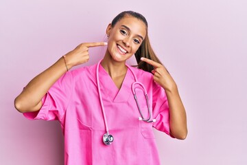 Young hispanic woman wearing doctor uniform and stethoscope smiling cheerful showing and pointing with fingers teeth and mouth. dental health concept.