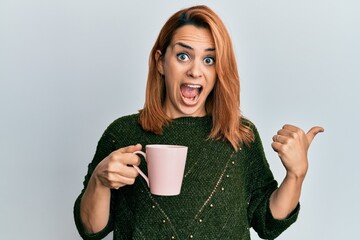 Hispanic young woman drinking a cup coffee pointing thumb up to the side smiling happy with open mouth