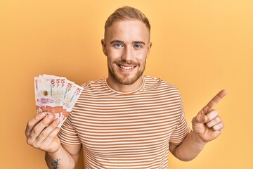 Young caucasian man holding 10 colombian pesos banknotes smiling happy pointing with hand and finger to the side