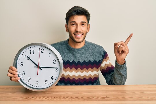 Young handsome man holding big clock smiling happy pointing with hand and finger to the side