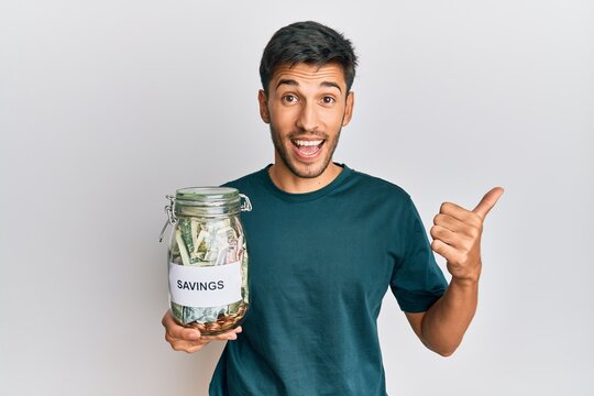Young handsome man holding jar with savings pointing thumb up to the side smiling happy with open mouth
