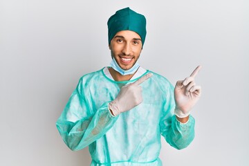 Young handsome man wearing surgeon uniform and medical mask smiling and looking at the camera pointing with two hands and fingers to the side.