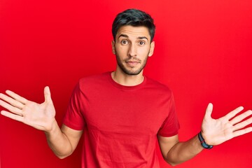 Naklejka premium Young handsome man wearing casual tshirt over red background clueless and confused with open arms, no idea and doubtful face.