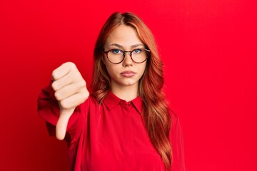 Young beautiful redhead woman wearing casual clothes and glasses over red background looking unhappy and angry showing rejection and negative with thumbs down gesture. bad expression. © Krakenimages.com