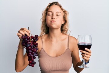 Beautiful caucasian woman holding branch of fresh grapes and red wine puffing cheeks with funny face. mouth inflated with air, catching air.