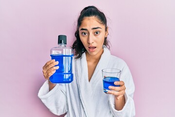 Hispanic teenager girl with dental braces holding mouthwash in shock face, looking skeptical and...