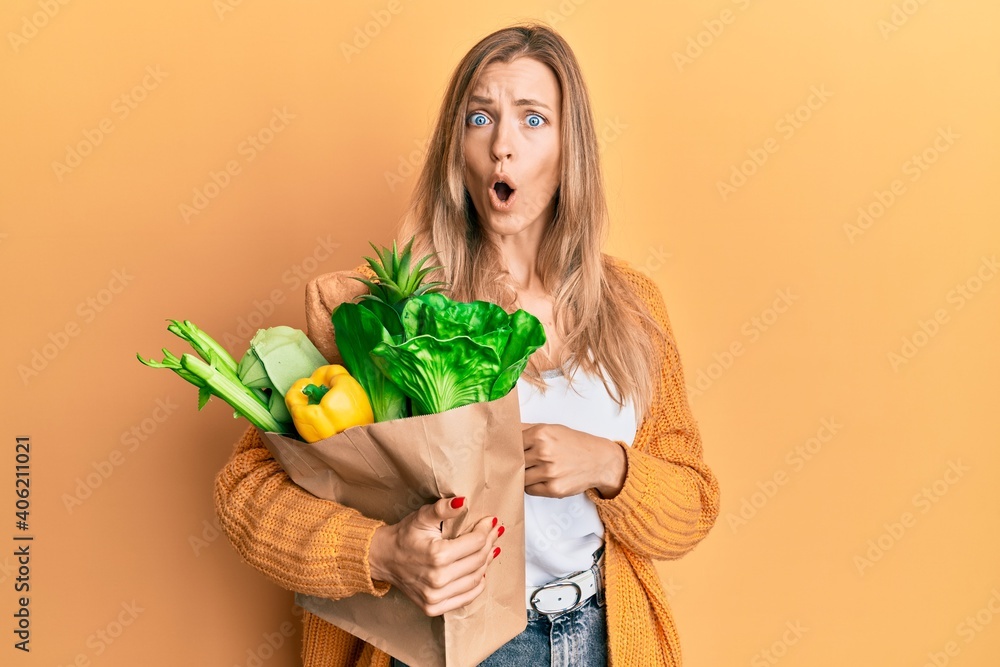 Sticker Beautiful caucasian woman holding paper bag with bread and groceries scared and amazed with open mouth for surprise, disbelief face