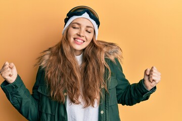 Beautiful blonde caucasian woman wearing snow wear and sky glasses very happy and excited doing winner gesture with arms raised, smiling and screaming for success. celebration concept.