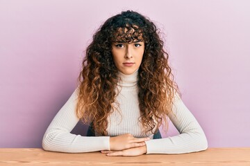 Young hispanic girl wearing casual clothes sitting on the table skeptic and nervous, frowning upset...