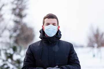 Fototapeta na wymiar A young guy in winter wears a mask. Young man in protective antiviral mask on the street. The guy in winter clothes in a protective mask. Young man in warm clothes and scarf on a winter day.