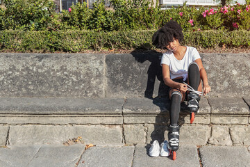 Young African American woman with afro hair is sitting putting on inline roller skates for outdoor...