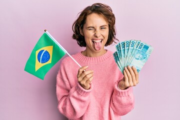 Young brunette woman holding brazil flag and real banknotes sticking tongue out happy with funny...