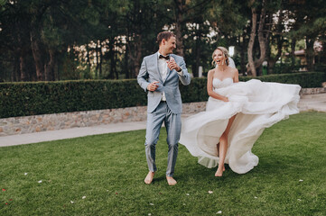 Naklejka na ściany i meble Stylish groom in a gray checkered suit and a blonde bride in a long white dress cheerfully and funny dance in a clearing with green grass, against the background of a forest barefoot.