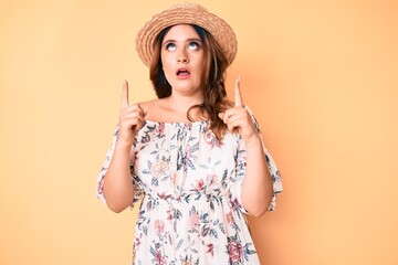 Young beautiful caucasian woman wearing summer dress and hat amazed and surprised looking up and...