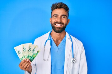 Young hispanic man wearing doctor uniform holding israel shekels looking positive and happy standing and smiling with a confident smile showing teeth