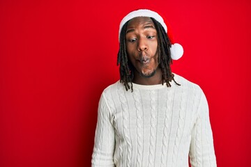 African american man with braids wearing christmas hat making fish face with lips, crazy and comical gesture. funny expression.