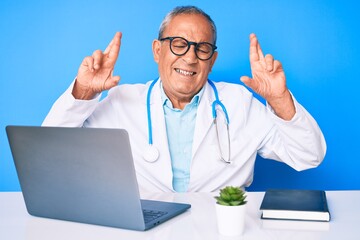 Senior handsome man with gray hair wearing doctor uniform working using computer laptop gesturing finger crossed smiling with hope and eyes closed. luck and superstitious concept.