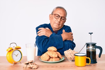 Senior handsome man with gray hair sitting on the table eating breakfast in the morning hugging...