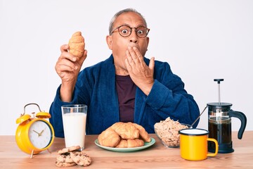 Senior handsome man with gray hair sitting on the table eating croissant for breakfast covering mouth with hand, shocked and afraid for mistake. surprised expression